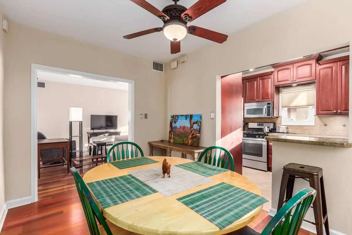 A round wooden table surrounded by green chairs is set in a dining area with a view extending into the adjacent kitchen. The decor is inviting and homey, offering a flexible space for shared meals, game nights, or quiet moments with coffee.