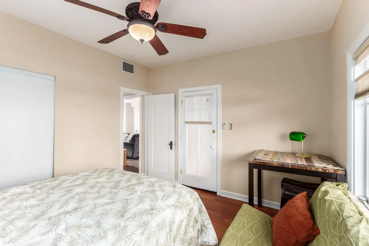 A serene bedroom with patterned bedding, a wooden table topped with a green lamp, and an open door providing a subtle glimpse of the outdoors. Soft natural light filters in, creating a cozy setting for relaxation, reading, or working at the table.