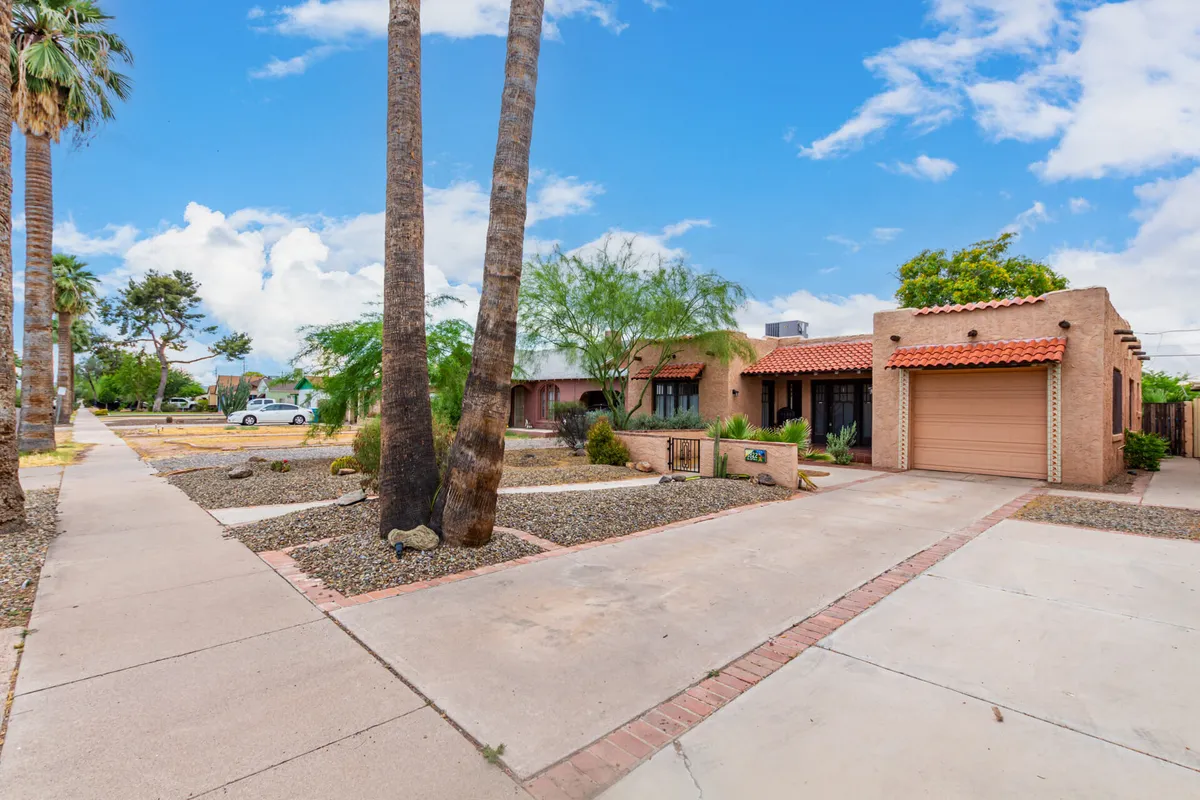 An inviting exterior view with palm trees, desert landscaping, a front porch, and a glimpse of the garage entrance. The setting is relaxed and neighborly, perfect for safe walks and enjoying the historic neighborhood atmosphere.