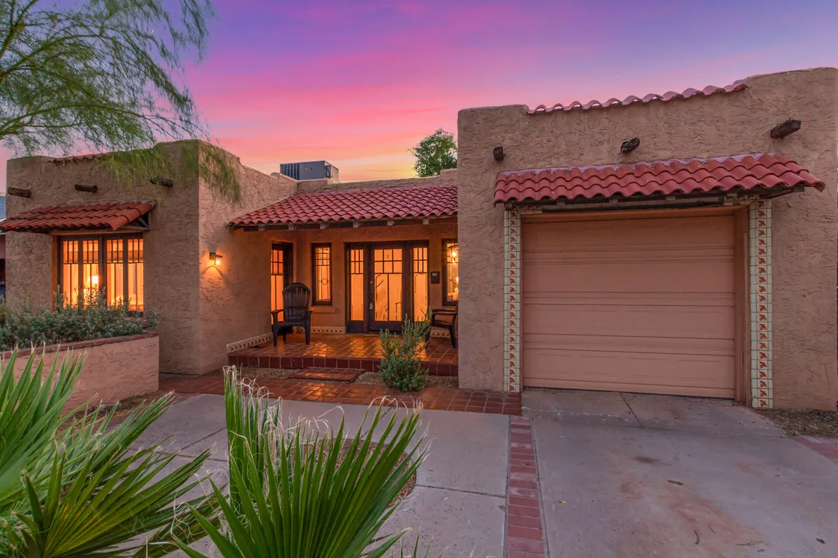 This image captures the charming front entrance of the property with double doors, adobe-textured walls, and a vibrant sunset sky overhead. The red-tiled roof and inviting porch area suggest a welcoming transition from home to the surrounding cityscape.